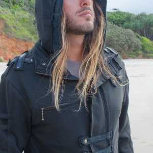 May include: Dark gray hooded jacket with a zipper closure and decorative straps. The jacket features a hood with a brim and is made of a thick, textured fabric. The jacket has a unique design with multiple pockets and decorative elements.