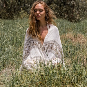 May include: A woman sits in a field of tall grass, wearing a white and brown patterned shawl. She has long brown hair and is wearing a silver necklace.