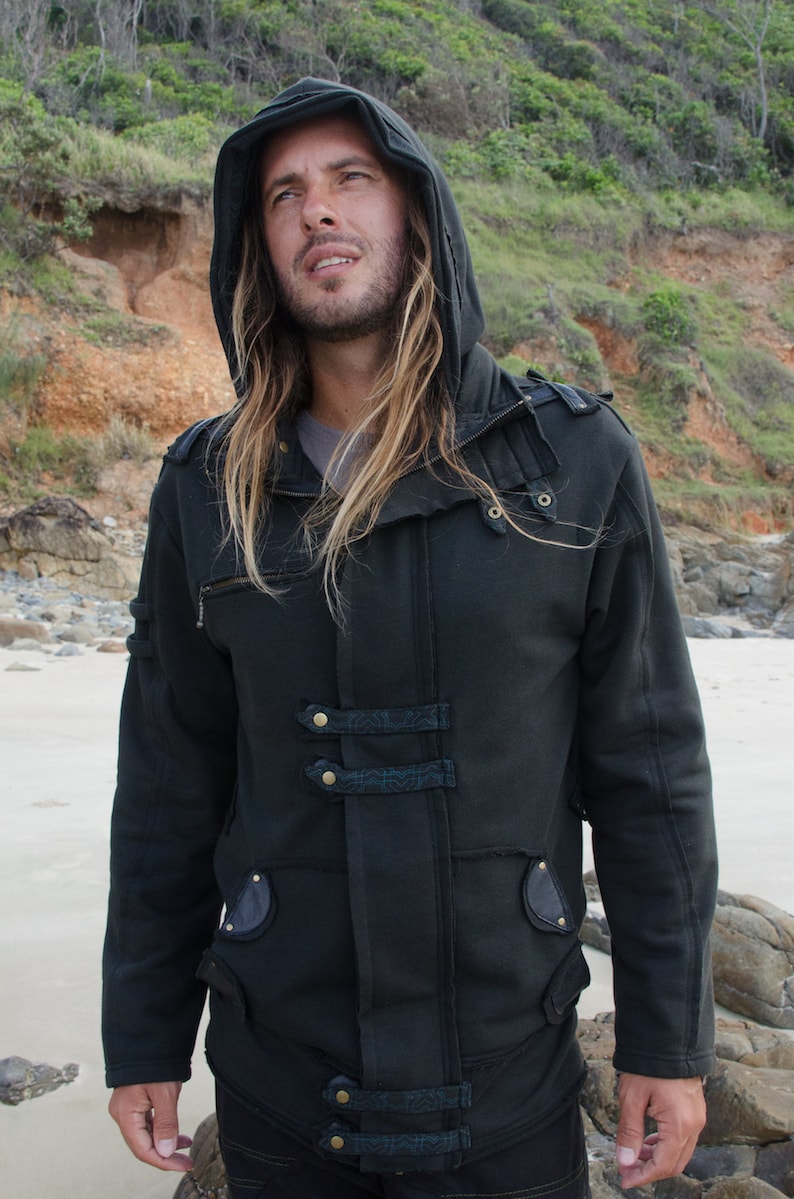 May include: A dark gray hooded jacket with multiple decorative straps and gold-colored buttons. The jacket has a zipper closure and pockets. The person in the image is wearing the jacket on a beach with a sandy shore and green foliage in the background.