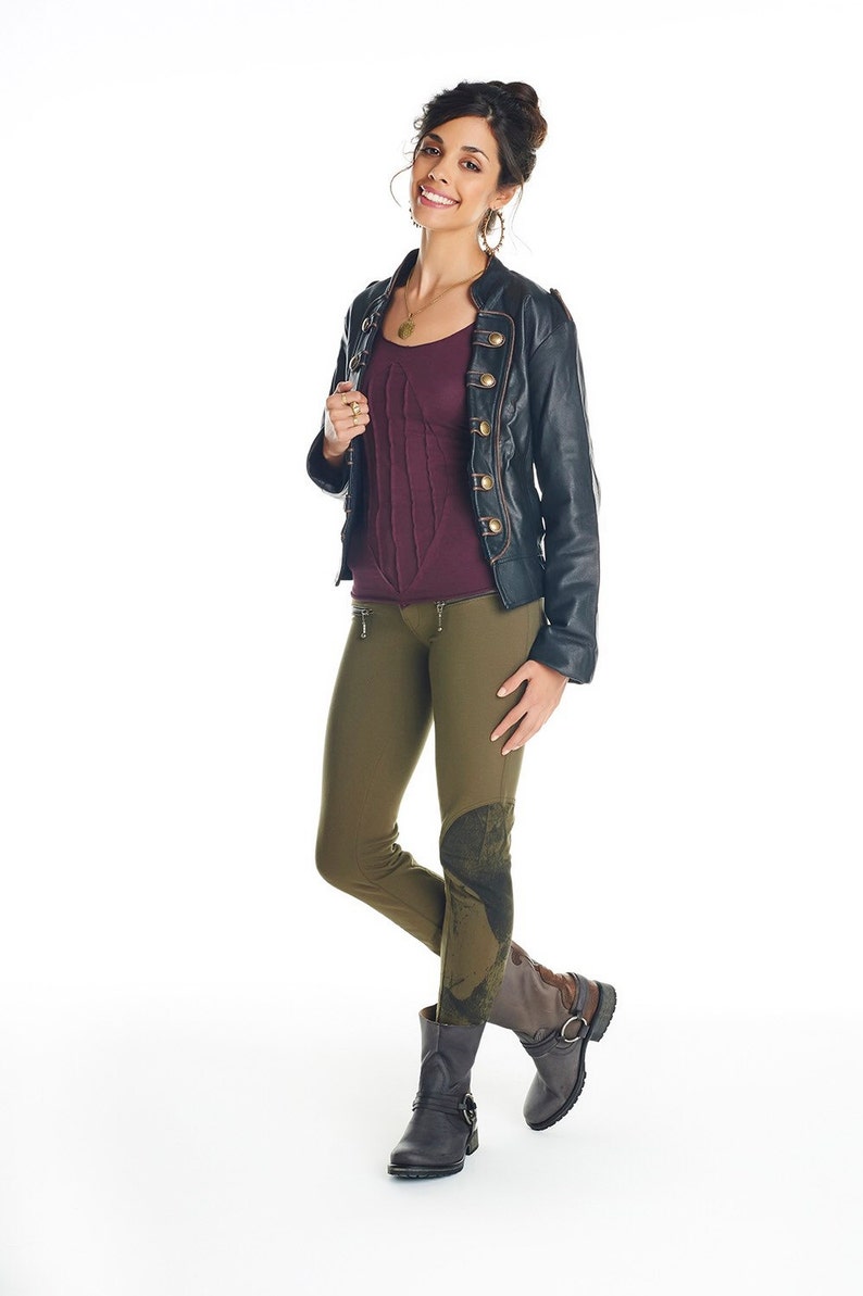 May include: A woman wearing a black leather jacket with gold buttons, a burgundy top, and olive green pants with a graphic print. She is wearing brown boots with buckles.