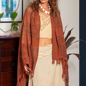May include: A woman wearing a rust-colored shawl with a geometric pattern and fringe. She is also wearing a beige crochet top and a white beaded necklace. The shawl is draped over her shoulders and the fringe is hanging down her back.