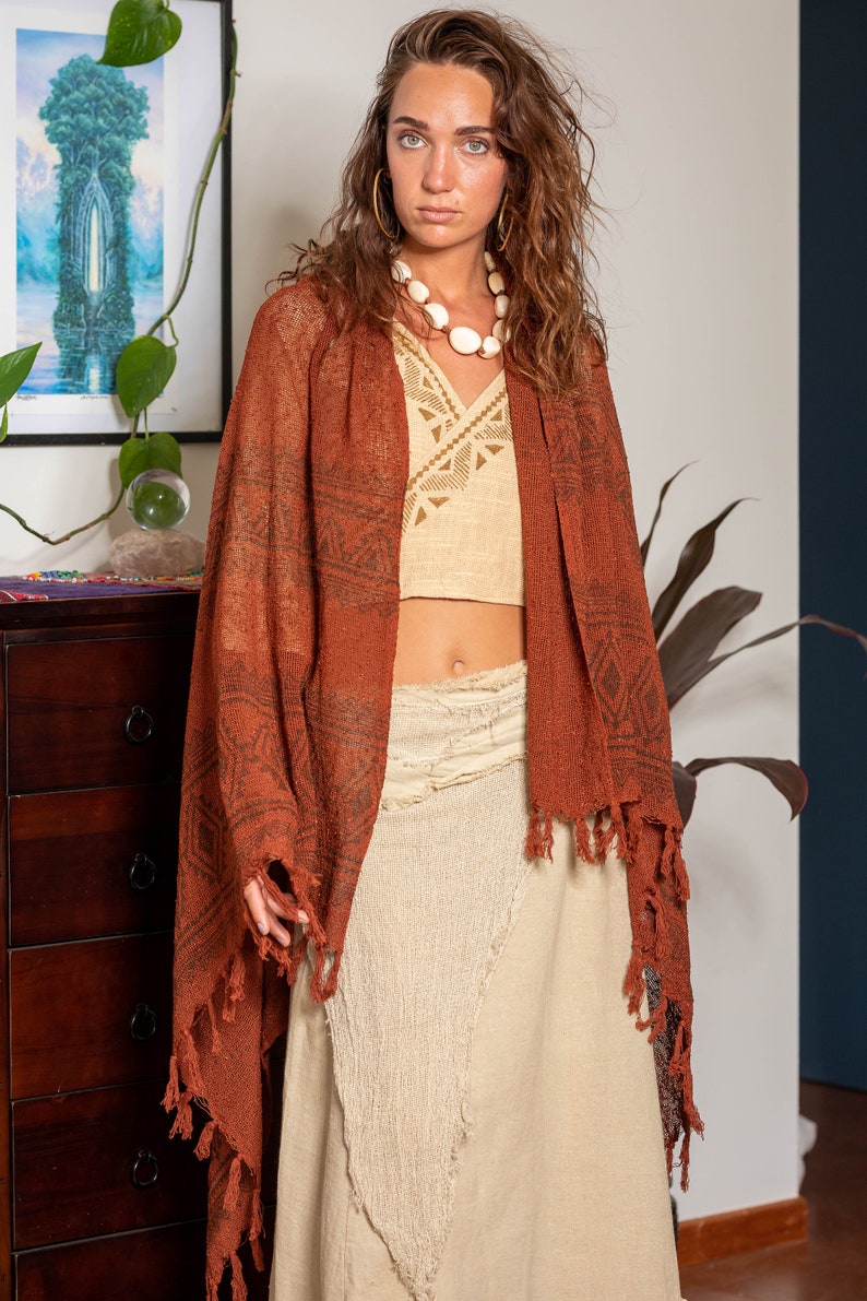 May include: A woman wearing a rust-colored shawl with a geometric pattern and fringe. She is also wearing a beige crochet top and a white beaded necklace. The shawl is draped over her shoulders and the fringe is hanging down her back.