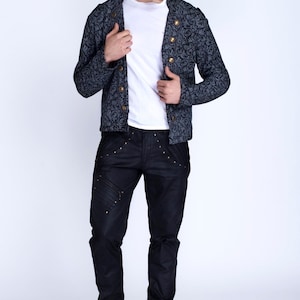Puede incluir: Un hombre con una chaqueta azul oscuro con estampado y botones dorados, pantalones vaqueros negros con costuras decorativas y zapatos negros.