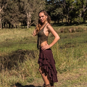 May include: A woman wearing a brown halter top and a long, burgundy skirt with ruffles. She is standing in a grassy field with trees in the background.