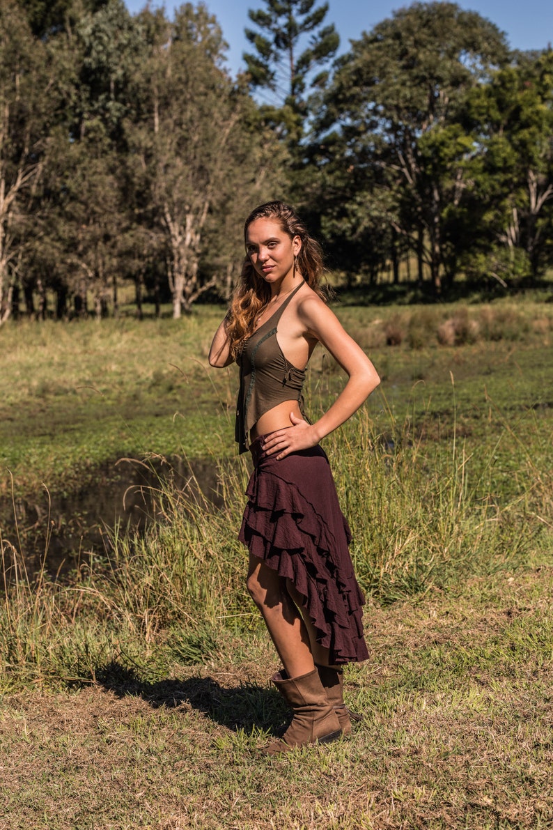 May include: A woman wearing a brown halter top and a long, burgundy skirt with ruffles. She is standing in a grassy field with trees in the background.