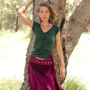 May include: A woman wearing a green, short-sleeved shirt with a burgundy skirt and a brown leather belt. She is standing next to a tree.