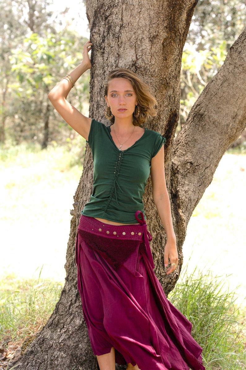 May include: A woman wearing a green, short-sleeved shirt with a burgundy skirt and a brown leather belt. She is standing next to a tree.