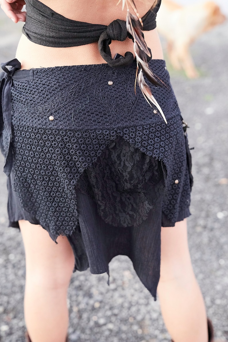 May include: Black crochet skirt with lace and fabric details. The skirt has a unique, asymmetrical design with a layered look. Embellished with small silver studs and feathers, it's a bohemian style piece.