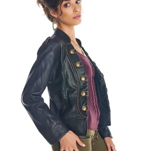 May include: A woman wearing a black leather jacket with gold buttons and brown trim, a burgundy shirt, and olive green pants. She is wearing gold rings on her fingers.