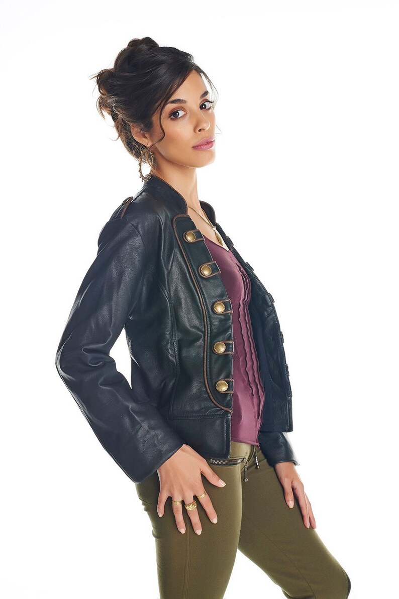 May include: A woman wearing a black leather jacket with gold buttons and brown trim, a burgundy shirt, and olive green pants. She is wearing gold rings on her fingers.