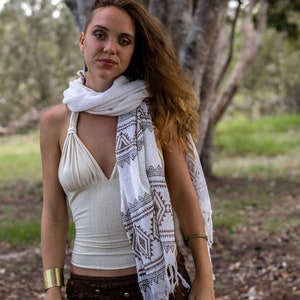 May include: A woman wearing a white and brown patterned scarf, a white halter top, and a brown skirt with fringe. She is standing in a forest setting.