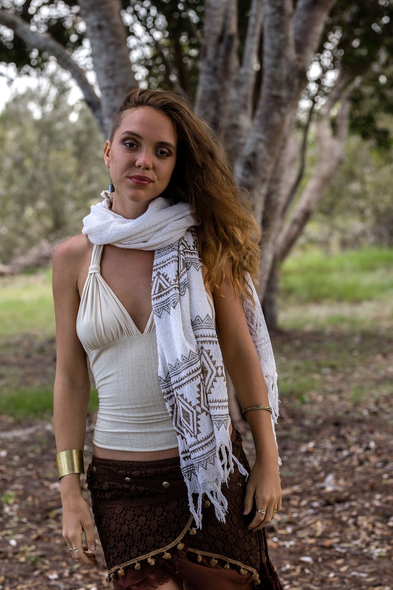 May include: A woman wearing a white and brown patterned scarf, a white halter top, and a brown skirt with fringe. She is standing in a forest setting.