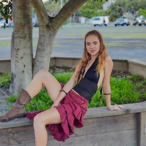 May include: A woman wearing a black halter top and a burgundy ruffled skirt sits on a wooden bench. She is wearing brown boots and has long brown hair.