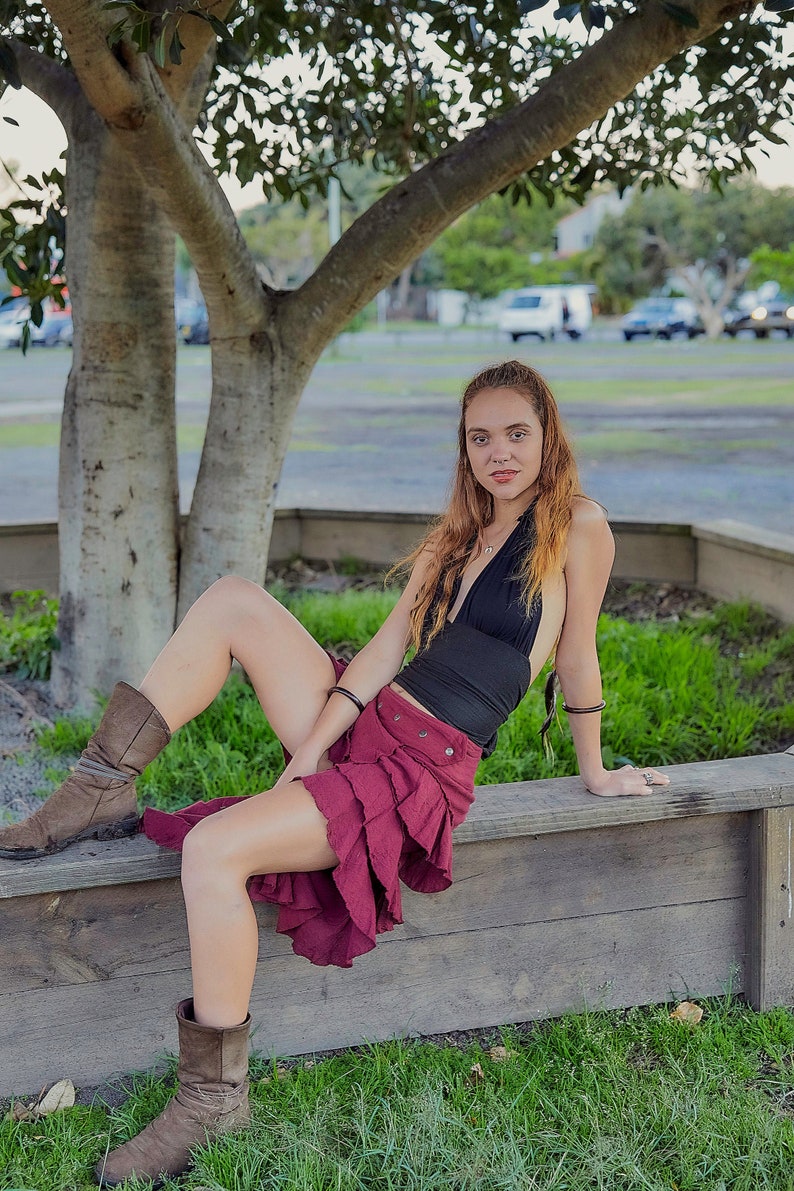 May include: A woman wearing a black halter top and a burgundy ruffled skirt sits on a wooden bench. She is wearing brown boots and has long brown hair.