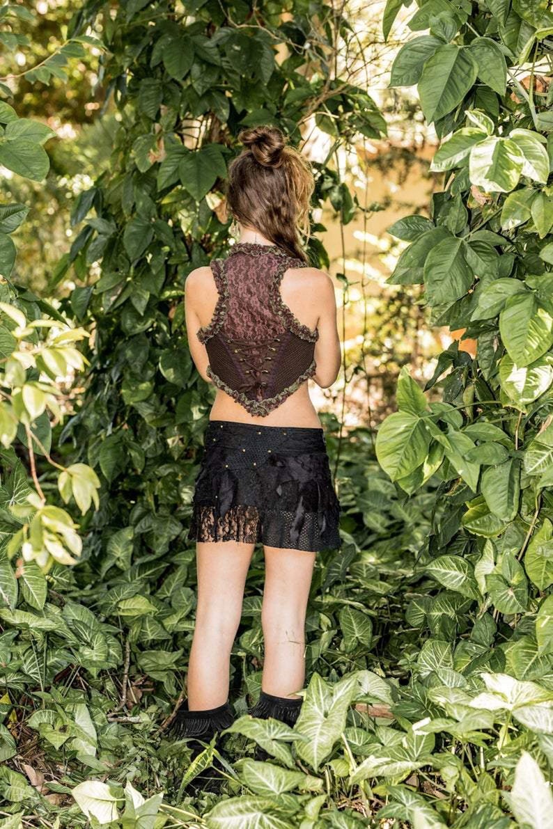 Pu&ograve; includere: Una donna indossa un gilet marrone e verde con una gonna nera e stivali neri. &Egrave; in piedi in un lussureggiante giardino verde.