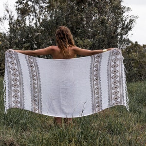 May include: A white cotton throw blanket with brown and gray geometric patterns. The blanket has fringe on the edges and is being held up by a person in a grassy field.