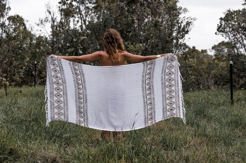 May include: A white cotton throw blanket with brown and gray geometric patterns. The blanket has fringe on the edges and is being held up by a person in a grassy field.