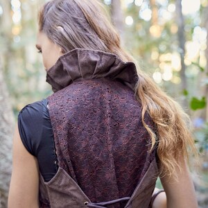 May include: A brown waistcoat featuring a high ruffled collar and a back panel made of dark brown floral lace. The garment includes a corset-style lace-up closure with metal eyelets at the lower back, layered over a plain black short-sleeved shirt.