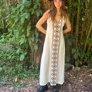 May include: A white maxi dress with a brown geometric pattern down the front. The dress has a V-neckline and a sleeveless design. The model is wearing a black hat and black shoes.