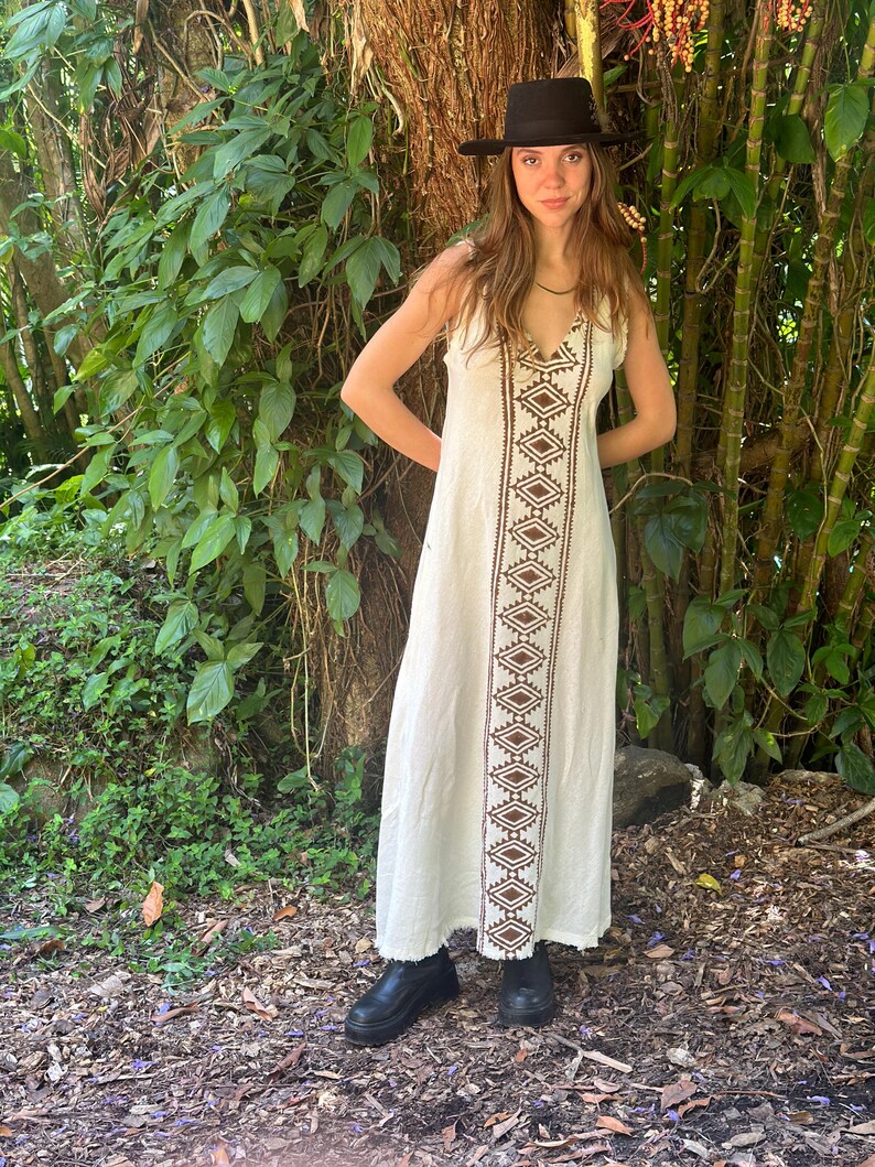 May include: A white maxi dress with a brown geometric pattern down the front. The dress has a V-neckline and a sleeveless design. The model is wearing a black hat and black shoes.