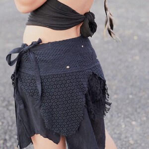 May include: Black wrap skirt with a crochet panel and fringed edges. The skirt features a tie closure and is paired with a black halter top. The outfit is accessorized with feathers and a wooden bracelet, creating a bohemian aesthetic.