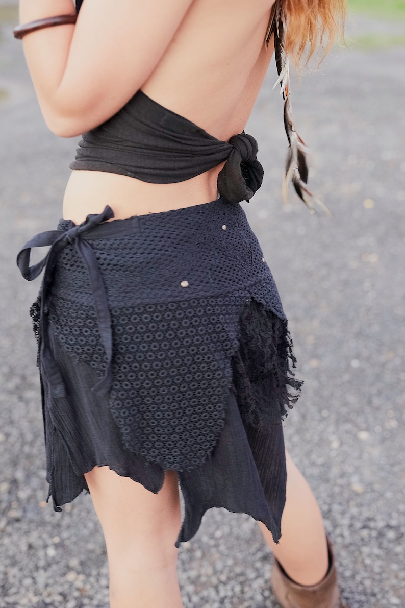 May include: Black wrap skirt with a crochet panel and fringed edges. The skirt features a tie closure and is paired with a black halter top. The outfit is accessorized with feathers and a wooden bracelet, creating a bohemian aesthetic.