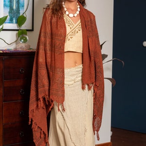May include: A woman wearing a long, rust-colored shawl with a geometric pattern and fringe. She is also wearing a white crop top and a long, white skirt with a frayed hem.