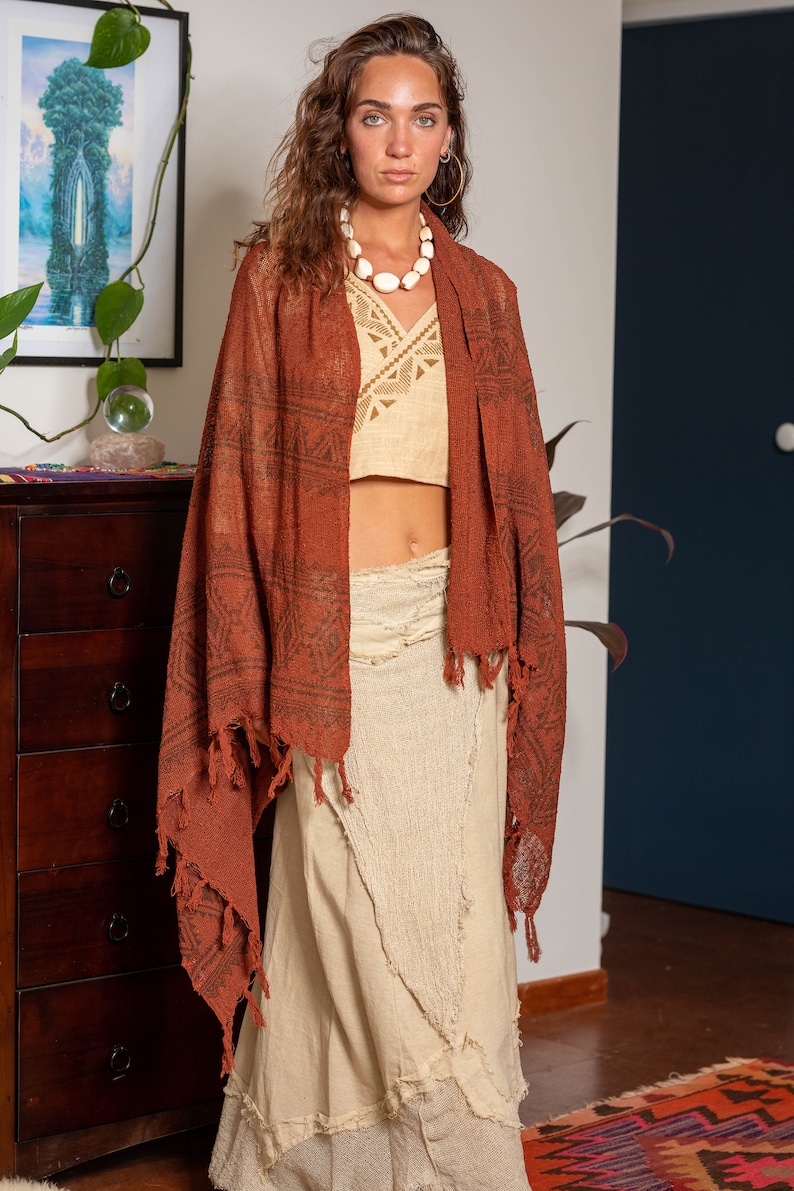 May include: A woman wearing a long, rust-colored shawl with a geometric pattern and fringe. She is also wearing a white crop top and a long, white skirt with a frayed hem.