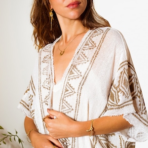 Puede incluir: Kimono blanco de crochet con estampado geom&eacute;trico marr&oacute;n. El kimono tiene cuello en V y mangas largas. La modelo lleva un collar y pendientes de oro.