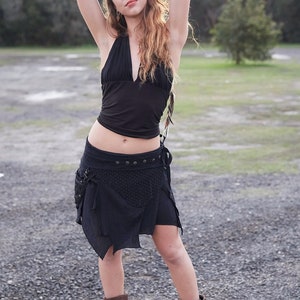 May include: A woman wearing a black halter top and a black wrap skirt with pockets. The skirt has a decorative pattern and is paired with brown boots. The background is a gravel path and trees.