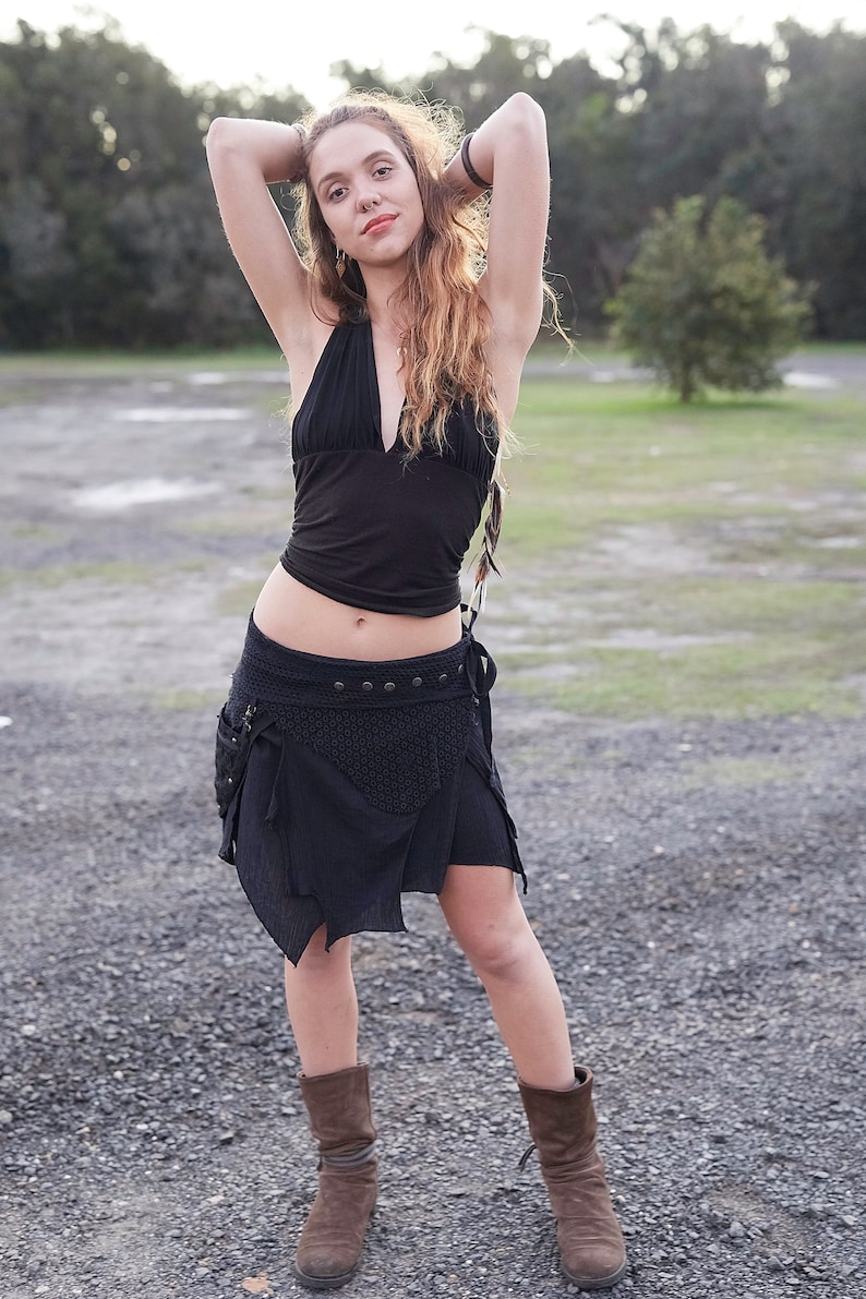May include: A woman wearing a black halter top and a black wrap skirt with pockets. The skirt has a decorative pattern and is paired with brown boots. The background is a gravel path and trees.