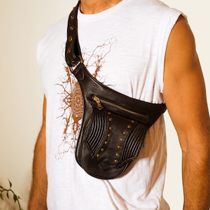 K&ouml;nnte beinhalten: Eine schwarze Leder-Umh&auml;ngetasche mit einem Rei&szlig;verschlussfach und Metallnieten. Die Tasche wird &uuml;ber der Schulter einer Person getragen, die ein wei&szlig;es T-Shirt tr&auml;gt.