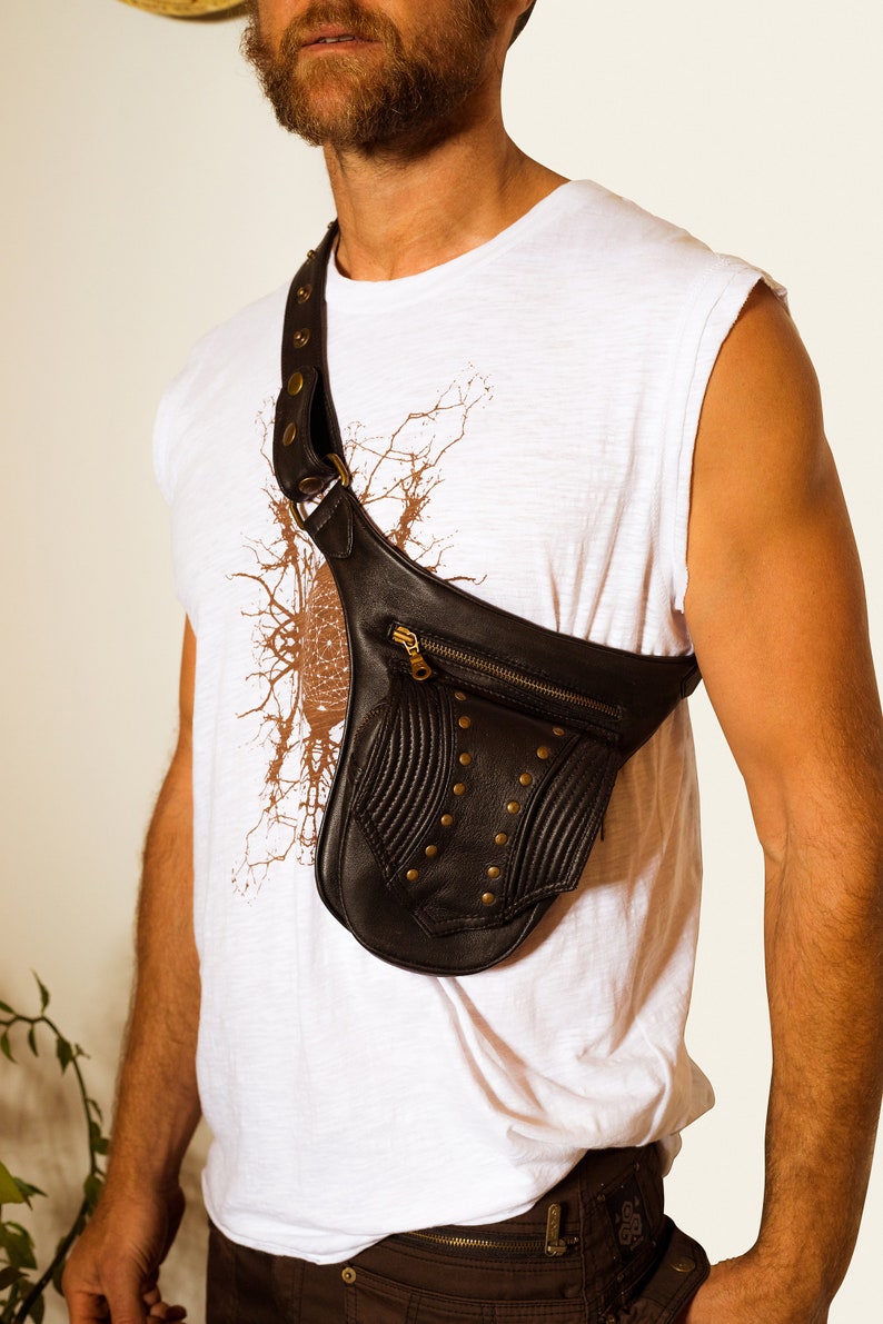 K&ouml;nnte beinhalten: Eine schwarze Leder-Umh&auml;ngetasche mit einem Rei&szlig;verschlussfach und Metallnieten. Die Tasche wird &uuml;ber der Schulter einer Person getragen, die ein wei&szlig;es T-Shirt tr&auml;gt.
