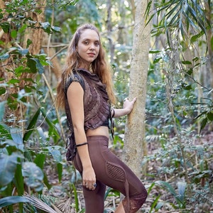 May include: A brown cropped vest with ruffled edges and metal studs paired with brown leggings featuring a leaf-shaped side cutout detail, lace trim at the hem, and brown boots. The outfit is worn in a natural outdoor setting with trees.