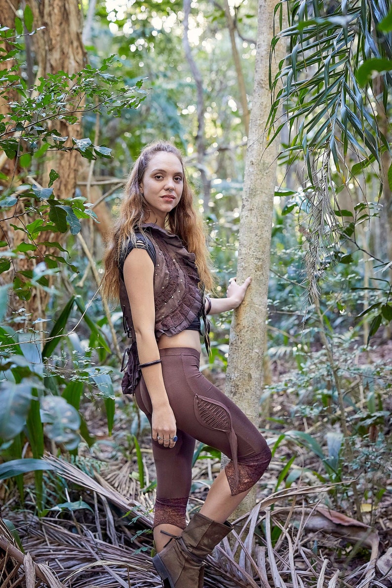 May include: A brown cropped vest with ruffled edges and metal studs paired with brown leggings featuring a leaf-shaped side cutout detail, lace trim at the hem, and brown boots. The outfit is worn in a natural outdoor setting with trees.