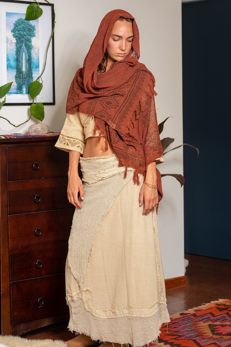 May include: A woman wearing a long, cream-colored skirt with a frayed hem and a rust-colored shawl with a geometric pattern.
