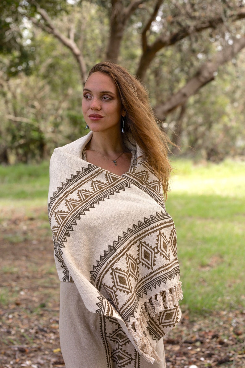 May include: A woman wearing a white shawl with a brown geometric pattern. The shawl has fringe on the edges. The woman is standing in a grassy field.