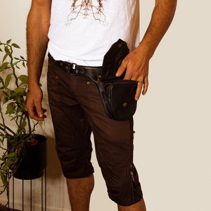 K&ouml;nnte beinhalten: Eine Person tr&auml;gt eine braune Shorts mit einer schwarzen Leder-Bauchtasche, die an der rechten H&uuml;fte getragen wird. Die Bauchtasche hat einen Rei&szlig;verschluss und einen Schnallenverschluss. Die Person tr&auml;gt auch ein wei&szlig;es &auml;rmelloses Shirt mit einem braunen grafischen Design.
