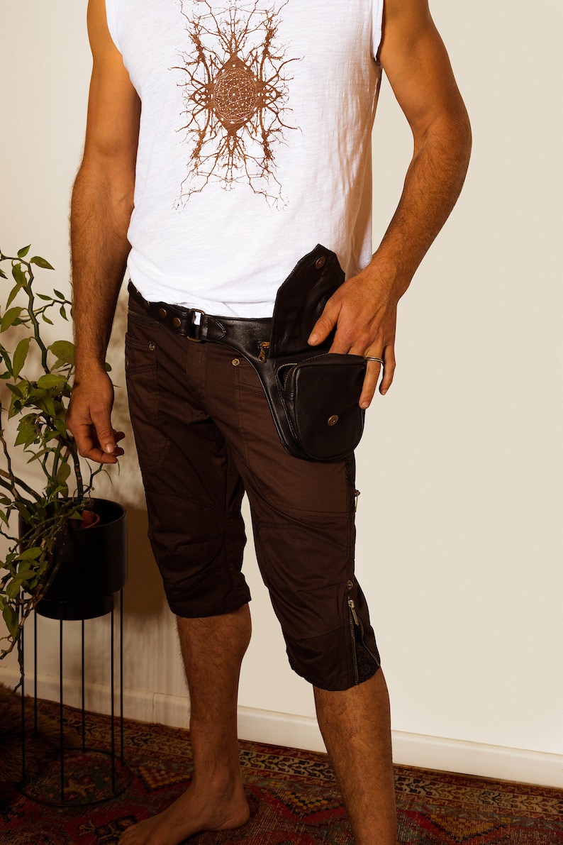 K&ouml;nnte beinhalten: Eine Person tr&auml;gt eine braune Shorts mit einer schwarzen Leder-Bauchtasche, die an der rechten H&uuml;fte getragen wird. Die Bauchtasche hat einen Rei&szlig;verschluss und einen Schnallenverschluss. Die Person tr&auml;gt auch ein wei&szlig;es &auml;rmelloses Shirt mit einem braunen grafischen Design.