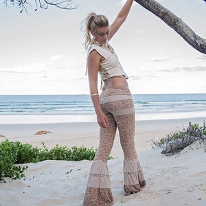 May include: A woman wearing a white crop top and brown bell bottom pants with lace trim. She is standing on a sandy beach with a tree branch overhead.
