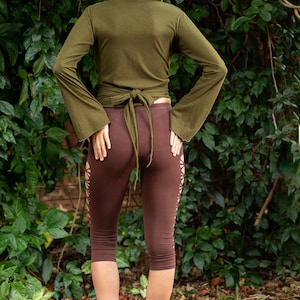 May include: A woman wearing a green hooded wrap top with a brown tie and brown leggings with cutouts. She is standing in front of a green leafy background.