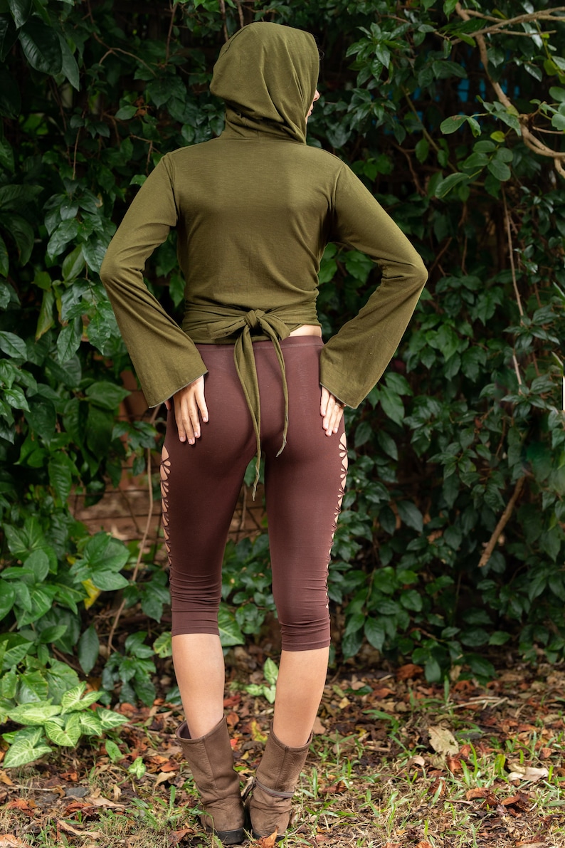May include: A woman wearing a green hooded wrap top with a brown tie and brown leggings with cutouts. She is standing in front of a green leafy background.