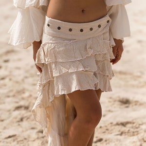 Puede incluir: Una falda blanca de volantes de varios niveles con un cinturón de tela marrón y detalles metálicos. La falda es asimétrica, con un lado más largo que el otro.