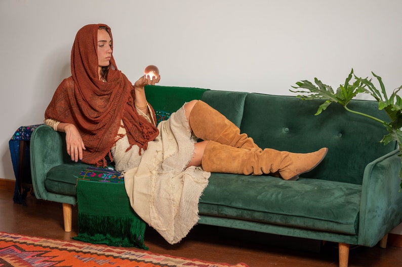 May include: A woman wearing a long, brown shawl and brown suede boots sits on a green velvet sofa. She is holding a crystal ball in her hand.