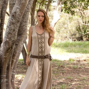 May include: A woman wearing a long, beige linen dress with brown geometric embroidery. She is standing in a forest setting with trees in the background. The dress has a brown leather belt with metal accents.
