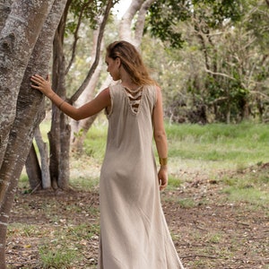 May include: A woman wearing a long, light brown linen dress with a unique criss-cross design on the back. She is standing in a forest setting, with trees and green grass in the background.