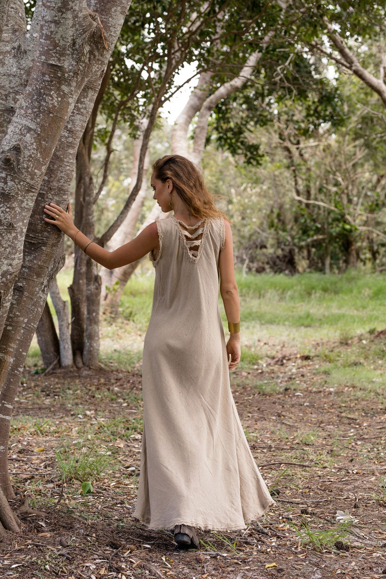 May include: A woman wearing a long, light brown linen dress with a unique criss-cross design on the back. She is standing in a forest setting, with trees and green grass in the background.