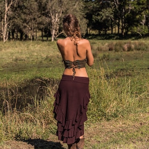 May include: A woman wearing a brown, ruffled skirt and a green, lace-up top. She is standing in a grassy field with trees in the background.