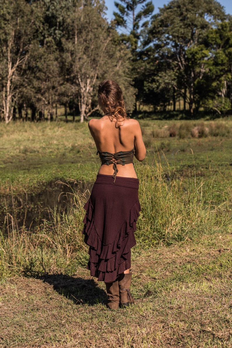 May include: A woman wearing a brown, ruffled skirt and a green, lace-up top. She is standing in a grassy field with trees in the background.