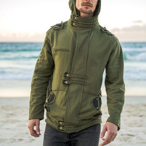 May include: An olive green hooded jacket with multiple button closures and decorative accents. The jacket features a high collar and a fitted silhouette. The person in the image is wearing dark gray jeans. The background is a beach with the ocean in the distance.